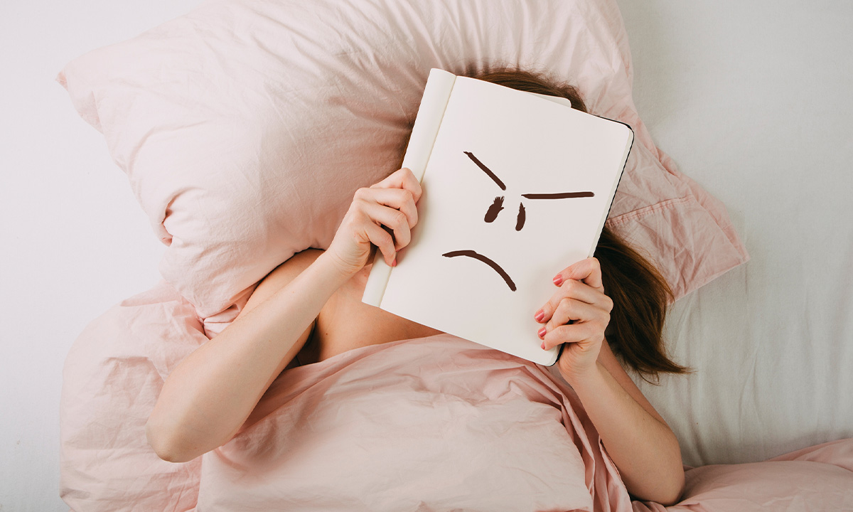 Im Bett liegende Frau hält sich eine Skizze eines mürrischen Gesichtsausdrucks vor das Gesicht