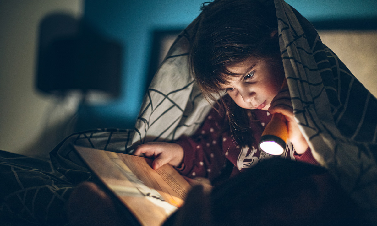 Junge sitzt unter Bettdecke mit Buch und Taschenlampe