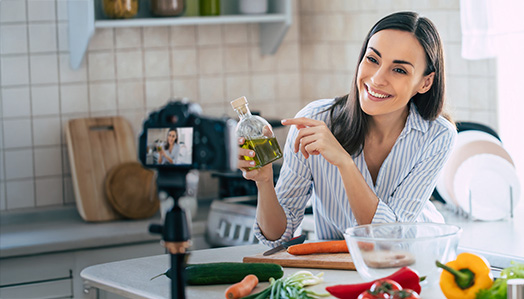 Frau an Küchentheke mit Gemüse zeigt auf Öl und zeichnet Video auf.